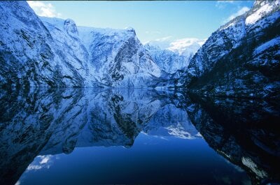 Bildet av fjord med snøkledde fjell på begge sider. Fjorden er stillestående og fjellene reflekteres i vannet